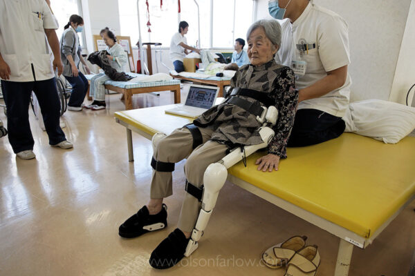 Japan’s Aging Population | Robotic Legs for Elderly
The Rehabilitation Section at the Akanekai Showa hospital uses these HAL legs to help elderly who have a hard time walking. These are in use on a daily basis. Outpatients (usually) come in twice a week and have a number of electrodes placed in key areas on their legs and lower torso. These electrodes monitor minor changes in muscle activity and transfer that information to the robotic joints. The woman being put into the legs is EMOTO, Chiyoko (89). She had fractures of her right and left thighbone. The main therapist working with her is TSURUGA, Mai (P.T.).
All of these robotics folks talk about studies showing that more and more elderly are isolated and have no one to relate to. In 2009, nineteen percent of Japan’s elderly died with no one around them. They have a word for it: “Kodokushi,” meaning “lonely death.”
