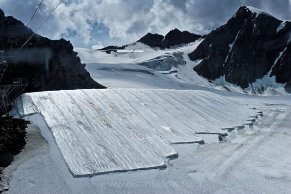 An attempt to keep snow from melting creates an austere, surreal scene at a ski run in Austria. The Alps are pristine at higher reaches, but the effects of climate change and global warming is seen at lower elevations.  Integral to the local economy, ski resorts must be protected from higher temperatures that melt the ice. Workers are hired to cover the snow pack with a fleece blanket to keep it from melting which seems equivalent to putting a band-aid on a glacier.
The Alpine glaciers — in Austria, Switzerland, France and Italy — are losing one percent of their mass every year and, even with no acceleration in that rate, they will have all but disappeared by the end of the century.
Photograph was published in “Meltdown, The Alps Under Pressure” in National Geographic magazine.
