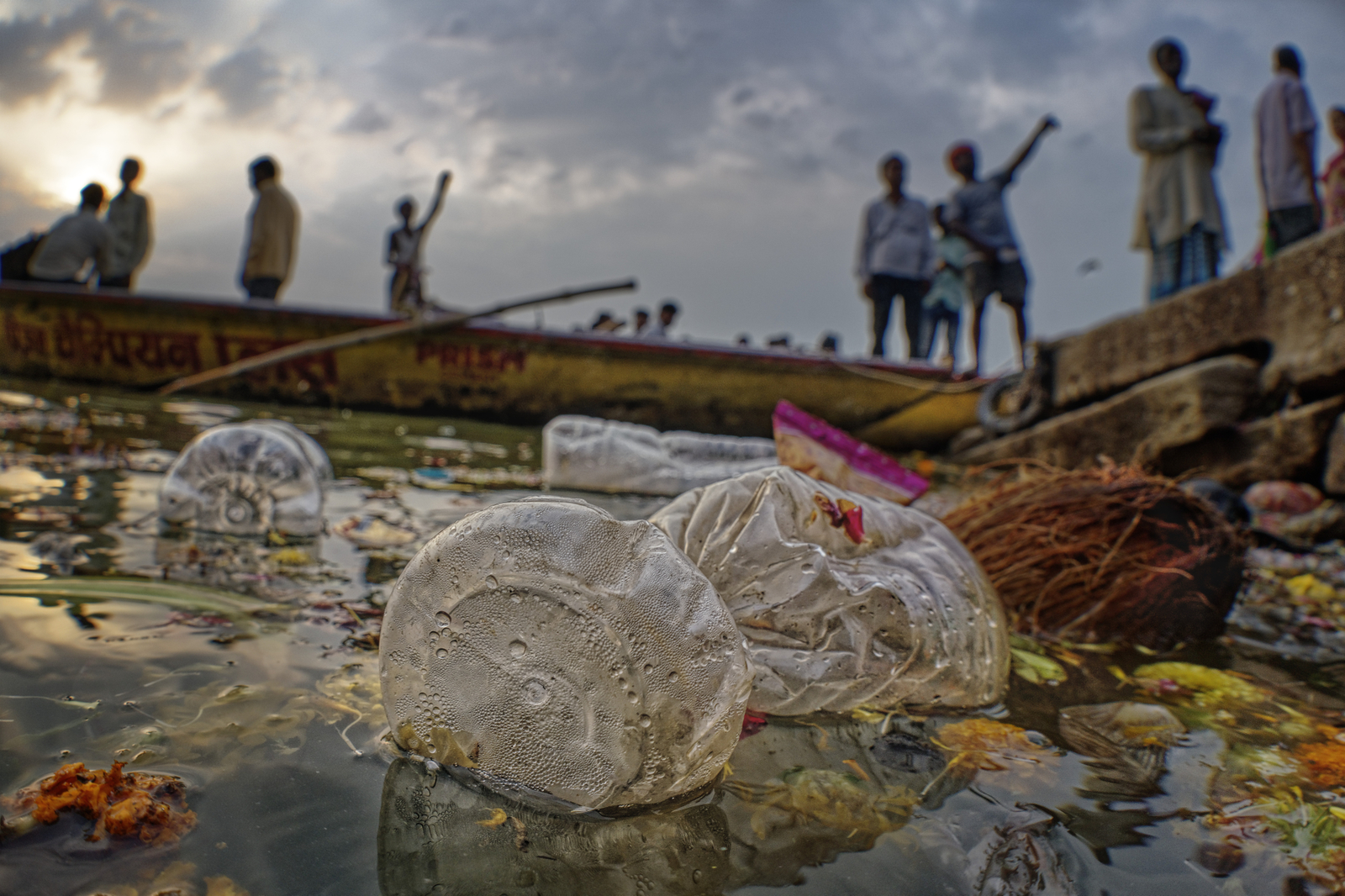 plastic bottles and flowers float in water with human figures in the background