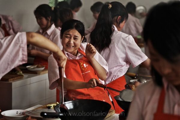 Girls from rural countryside learn to be maids for the newly wealthy class. They learn to cook and iron at the Fuping Vocational Skills Training School. Li Lingping responds to flying grease in one of the cooking classes.
Since opening up its economy in 1978 and moving toward a market economy, China has lifted about 400 million people out of poverty, according to the World Bank. But this has led to wide income inequalities that the Communist Party is trying to address through its notion of a “harmonious society” that has a more even distribution of the benefits of recent decades of speedy economic growth. Migrant workers in China are mostly people from impoverished regions who go to more urban and prosperous coastal regions in search of work. According to Chinese government statistics, the current number of migrant workers in China is estimated at 120 million (approximately 9% of the population). China is now experiencing the largest mass migration of people from the countryside to the city in history. An estimated 230 million Chinese (2010), roughly equivalent to two-thirds the population of the U.S., have left the countryside and migrated to the cities in recent years. About 13 million more join them every year—an expected 250 million by 2012, and 300 to perhaps 400 million by 2025. Many are farmers and farm workers made obsolete by modern farming practices and factory workers who have been laid off from inefficient state-run factories. Men often get construction jobs while women work in cheap-labor factories. So many migrants leave their homes looking for work they overburden the rail system. In the Hunan province, 52 people were trampled to death in the late 1990s when 10,000 migrants were herded onto a freight train. To stem the flow of migrants, officials in Hunan and Sichuan have placed restrictions on the use of trains and buses by rural people. In some cities, the migrants almost outnumber the residents. One young girl told National Geographic, “All the young people leave our village. I’m not going back. Many can’t even afford a bus ticket and hitchhike to Beijing.” Overall, the Chinese government has tacitly supported migration as means of transforming China from a rural-based economy to an urban-based one.
