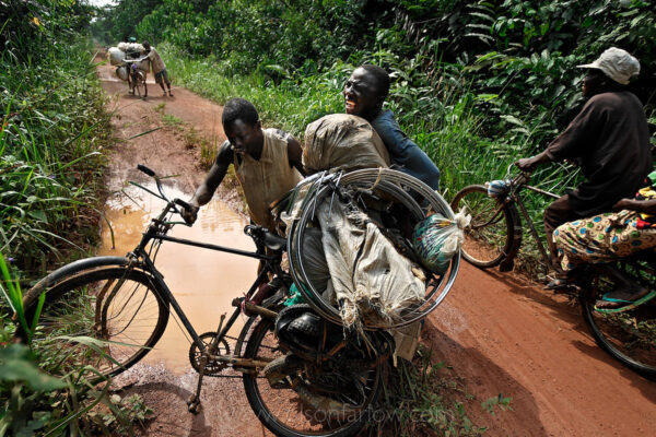Human Flood From Albertine Rift Seeking Gold | Ituri, Congo
A human flood chokes the mud-slick remnants of a highway, the main vein for commerce through the Ituri. Rough and rutted during dry weather, the roads are nearly impassable in the wet season. But rain can’t stop the toleka traders, who push goods hundreds of miles by bicycle to resupply the newly minted gold mines in the area.
