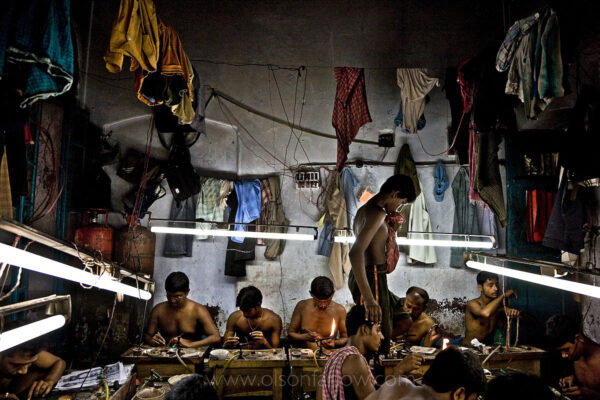 Artisanal Gold Smiths Sweat Shop | Kolkata (Calcutta), India
These guys make, at best, about 3-400 $USD per month. They live in the workplace. The clothes hanging above them are their clothes, their closet as it were.  At night they move the worktables aside and sleep on the floor in the same small space they work in. These guys are all very kind to each other; they are from the same village. They take turns going out for food, for tea. They take care of each other and they need each other. When one of them goes back to the village, he carries the money from all of them. When he comes back he brings things for everyone else from all of their families.
The goldsmiths are given 102.6 grams of gold and are expected to return 100 grams of jewelry. The average waste for 100 grams is 1.3 grams.  So the owner of the factory tells them he wants a 20-gram piece of jewelry and they have to figure out how to make it that weight and keep to the design they are given.
