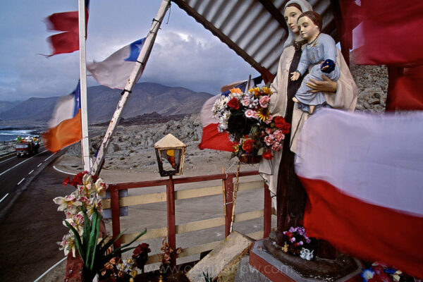 Colorful flags, religious statuary and plastic flowers decorate an alter that marks the Pan American highway in a wind-blown desolate section of the Atacama Desert in northern Chile.  Animitas,are the name of the roadside shrines to the dead that line the roads, numbering in the tens of thousands, especially along rural highways and in poor and working-class areas of small cities and towns. The word refers not only to the shrine, but to the soul of the dead housed there. Animitas are not simply memorial markers, but serve as homes for the souls of the departed. They are places where the living can visit and seek intercession from those souls, the suddenness of whose death gives those deceased special spiritual power. Bodies are not buried there. Instead, the animitas mark the spot where body and soul were separated, and where the soul may linger and still be reached, at least for a while, by the living. According to a Catholic writer, they are not sanctioned by the church.
