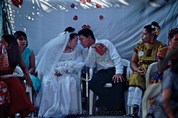 A Zapotec bride and groom share a private moment at their wedding party. Dancing and celebrating has begun under a tent the covers the  guests and festivities. Tradition rules that the man’s side of the family covers the cost of the mass, food, drinks, band–everything connected with the wedding and party. The woman’s family gives money and presents to the couple. The party can last up to three or four days with the total cost hovering around $20,000 U.S.
