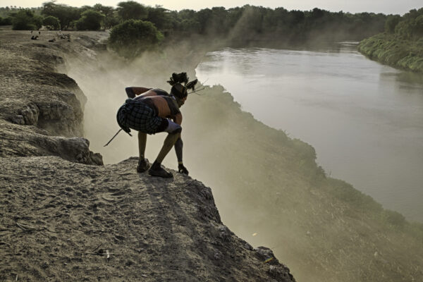 An elder of the Kara tribe watches his goats at dusk peering out over the bank of the Omo River. His tribe once controlled land on both sides of the river, but an enemy tribe gradually encroached on their territory. Before the peace agreement, a member of the warring tribe could have killed him if he was exposed in this way.  The Kara survive on growing sorghum on the banks and their animals.
In the background is the river that flows south toward Lake Turkana. The Omo River is 760 kilometers long and starts in the Shewan Highlands and empties into the lake. The coverage area for this story is located in the region between the Gibe III Dam in the north to the first bridge ever built over the Omo, at Omorate, near the Kenyan border.
