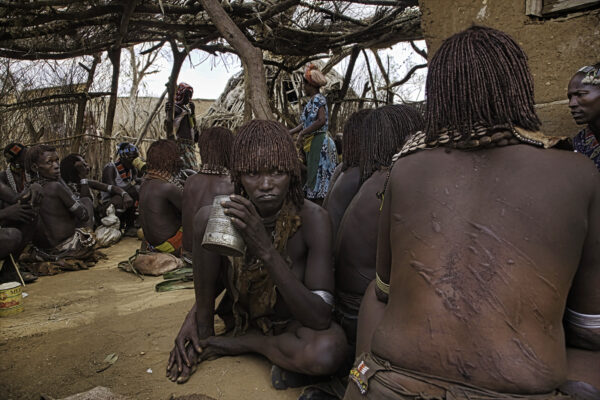 Women wait for a bull-jumping ceremony to begin in a local bar in Turmi, a Hamar town in the Omo River Valley. The scars on the their backs are from participating in a previous bull jumping ceremony where they are whipped. The marks are a sign that the women are supported by the community in general, but more specifically, from the boys that became men during the bull jumping ceremony. They are drinking beer made from food aid sorghum that was subsequently fermented for alcohol. 
