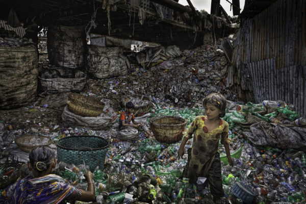 



A woman works sorting while her daughter wades through a sea of plastic under the Buriganga Bridge in Dhaka, Bangladesh. This family is part of informal plastic waste industry and set up their operation working long hours to eke out of living looking for recyclable materials.  It may appear a chaotic, tangled heap but the workers make order finding like colors and types in the waste that is in the shadows of Burigonga Bridge Road that goes over a backwater to the Buriganga River.




