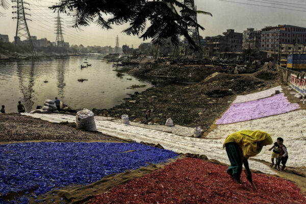 



Colorful bottles caps create piles of drying plastic  along a riverbank. Families wash shredded plastic for profit organizing it by color for recycling in Bangladesh’s informal plastic waste industry. Their hand labor is more accurate than highly industrialized recycling in the USA and the labor costs $2-$4 a day.  Blue bottle caps are sorted from red bottle caps and they are sorted from the green bottle caps. A huge overburden of plastic is thrown away landing in the river and washing out into the Bay of Bengal.




