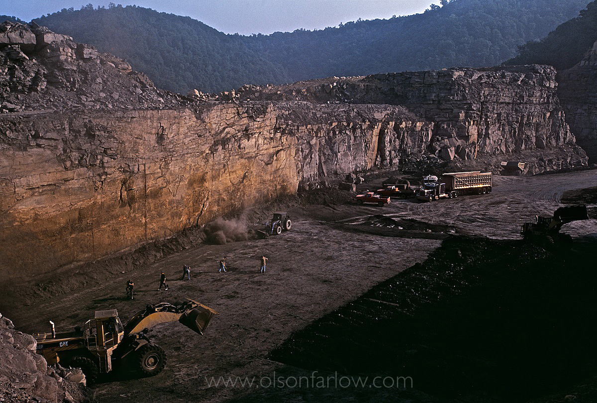 Bulldozer in pit mine with truck in background and small workers on the ground.