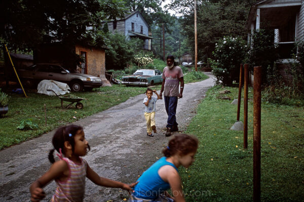 A man walks down the road in Tom Biggs Hollow in Letcher County, Kentucky, while his children play nearby.
Lucious Thompson, who lives in nearby Tom Biggs Hollow, joined Kentuckians for the Commonwealth when he found his land disrupted from above. “There’s good mining and there’s bad mining,” Mr. Thompson said. “Mountaintop removal takes the coal quick, 24 hours every day, making my streams disappear, with the blasting knocking a person out of bed and the giant ‘dozers beep-beeping all night so you cannot sleep.”
Mr. Thompson spoke with the authority of a retired underground miner. Underground miners led quieter, more pastoral lives above harsh, deep workplaces that were far out of sight. Now, the hollow dwellers have become witnesses more than miners as a fast-moving, high-volume process uses mammoth machinery to decapitate the coal-rich hills. 
“They make monster funnels of our villages,” said Carroll Smith, judge-executive, the top elected official, here in Letcher County, the location of some of the worst flooded hollows adjoining mountaintop removal sites. “They haven’t been a real good neighbor at all.”
With underground mining, coal miners led quieter, more pastoral lives above harsh workplaces deep in the ground and far out of sight. With mountaintop removal, a fast, high-volume process that uses mammoth machinery to decapitate the coal-rich hills that help define the hollows, the residents have become witnesses more than miners.
New York Times, http://www.nytimes.com/2002/08/11/national/11MINE.html
 
 
