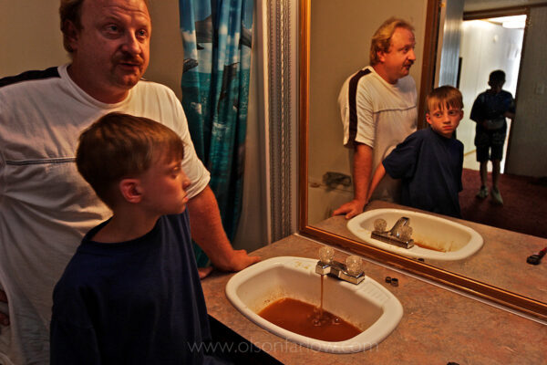 A father and sons react to the orange/black water in the bathroom that smells bad and has an oily residue mixed with coal soot. His health is affected and he fears for his sons. He suffers from rashes and red eyes when he showers and he tells the kids, “Don’t brush teeth in water. Don’t drink the water.”
He told National Geographic that when he moved in six years ago he thought the only problem was that the water was discolored by iron. At 38 years old, he has since developed gallstones, breathing problems, memory loss, and his hair is falling out. He has anxiety, nervousness, and his pancreas is at two percent function. All of this occurred after he moved to this trailer. Scared for his family, he asks, “What have I done to them?” Neighbors in nearby Williamson and Rawls who have the same water are having it analyzed.
A November 4, 2003 Associated Press article by Michelle Saxton of the Williamson Daily News entitled “Water in Mingo Communities Contains Manganese” stated that some security guards quit opening valves on Massey pumps when they realized they were poisoning the community. In a later court hearing it was shown that Massey Coal Company had, indeed, Illegally injected slurry from the Rawls Sales Processing Company (Massey Coal Company subsidiary) impoundment into abandoned underground mines for at least eight years.
As of the fall of 2011, some 500 West Virginia residents are still in limbo over a suit brought against Massey energy over claims that it and Rawls Sales poisoned hundreds of drinking water wells with coal slurry.

