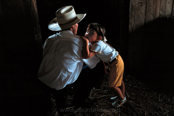 A father dons a white hat and stoops to watch horses with his daughter in the barn located in central Illinois along the Old National Highway.
