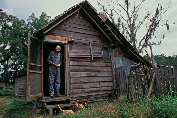 Elderly bachelor William McKinley Crews looks out the back door of his home near the Okefenokee National Wildlife Refuge. A new bull he bought earlier in the day broke through a fence and ran away, and he has waning hopes that it will return.
Crews led a reclusive life on his 160-acre farm in Moccasin Swamp, which borders the Okefenokee. His only company for many years was his older brother Daniel who was also a bachelor. Both were known for their skepticism of the outside world. Sought out by a reporter from the Miami Herald in 1982, the brothers were quoted as only fearing “God, the devil, women and electricity.”
Having no running water, electricity or telephone after his brother’s death, Crews lived mostly in isolation with his fourteen cats and four heifers he jokingly called his “nuns.”
