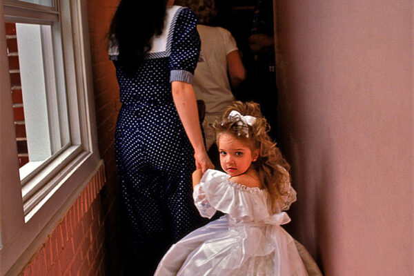 A young girl dressed in a long gown follows her mother down the hallway to the stage to compete in the beauty pageant honoring Mr. and Miss St. George, Florida. Beauty pageants became part of American society in the 1920s, and children’s pageants in the ‘60s.

