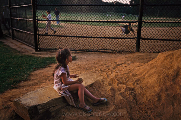 A young girl holds a baseball while watching older boys play in a ball field in Brooklyn’s Prospect Park.
In the original plan of the park, Frederick Law Olmsted, who designed with Calvert Vaux, appreciated pastoral scenery and long meadows broken only by clumps of deciduous trees. Those open views have been difficult to preserve in present day times as a demand for active recreational facilities has mounted in urban areas. The ball fields in Prospect Park were moved to the ends of the meadow, pushing the backstops to the edges to conform to Olmsted’s original plan.
