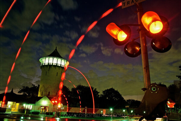 Flashing lights of a railroad crossing light the night sky in front of the water tower on central village square in Riverside, Illinois. Riverside is the first planned community in the United States, and was commissioned for a design by well-known landscape architect Frederick Law Olmsted and his partner Calvert Vaux. An affluent suburban community nine miles west of Chicago, Riverside maintains the original aesthetic charm that was planned to appeal to people desiring a “rural” location.
The town might not have ever been popular had it not been for the disastrous Chicago fire of 1871 which served as an impetus for people to move away from the crowded, urban setting.
In 1868, an eastern businessman named Emery E. Childs formed the Riverside Improvement Company, and purchased a 1,600-acre tract of property along the Des Plaines River and the Chicago, Burlington & Quincy Railroad line. The site was highly desirable due to its natural oak-hickory forest and its proximity to the Chicago Loop.
In 1869, the town’s plan called for curvilinear streets that followed the land’s contours and the winding Des Plaines River. The plan included a central village square, located at the main railroad station, and a Grand Park system that uses several large parks as a foundation, with forty-one smaller triangular parks and plazas located at intersections throughout town to provide additional green spaces.
