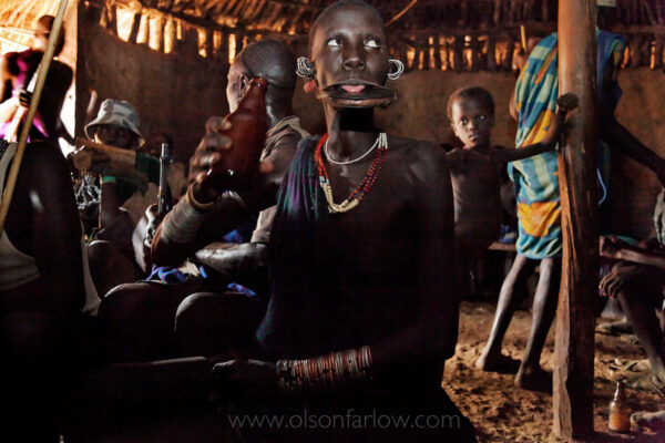 A Mursi woman licks the last drop of beer off of her lip plate in a bar in Kibbish which is the last village at the end of the last road in the upper Omo Valley.  Years of stretching a chin incision that was made during teenage years allows this woman to wear a lip plate.  The origin of this tradition is debated, but current belief is that it is purely for adornment. 
Women do not wear their lip plates while in their home village, so when they are in remote areas of Ethiopia only accessible by boat on the Omo River, they are free to wander their villages feeling safe with their lip plates off. Without an inserted plate, a ring of flesh hangs across their chins. The bar is walled in mud, its floor a cement of spit, sweat, and old bottle caps. Outside the bar, police walk up and down the town’s main dirt road with megaphones telling men to check their AK-47’s at the police station.  The man facing away in the background of this photo ignored their message.
