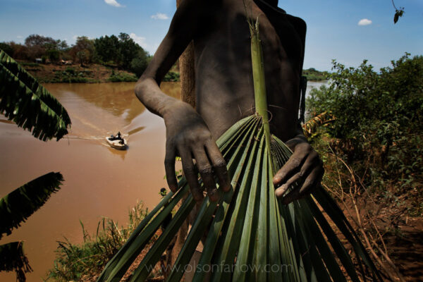 On the banks of the muddy Omo River, a Mursi child holds a leaf frond which is used as a chair.  This area of Ethiopia is isolated and the boat in the background is the only one on the river during an extensive six-week transect that went from the highlands, the origin of the river, to Kenya where the river flows into Lake Turkana.
