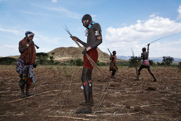 Checking the flex of his whips, a young Banna man prepares to deliver blows to a young woman during an initiation ritual, while in the background his counterpart winds up to whip another woman. Among the Ethiopian Banna and Hamar tribes in the Omo River Valley, it is customary for women to be whipped during the ceremony to initiate a boy into manhood. Female relatives of the initiate play a crucial supporting role—singing, dancing, and preparing food. The scars resulting from the whipping are a mark of pride for the women, showing solidarity with the boy who is enduring his own trials to become a man. Women sometimes blow small horns and sing songs, taunting the whippers and urging them to strike harder.  It is a symbol of belonging, and they are supported in the social structure of the Hamar, Karo and Bene tribes.
