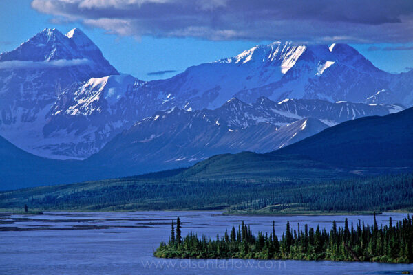 Snow-capped peaks of the Alaska Range rise beyond the Maclaren River north of the Denali highway.
Several peaks in view have elevations greater than 12,000 feet. Layers of snow accumulate and are compacted into ice, forming a glacier. As the glacier becomes heavier it moves down the slope, gouging the rock below. This glacial erosion contributes to the rugged, jagged appearance of the Alaska Range, and creates the long U-shaped valleys seen from the road.
