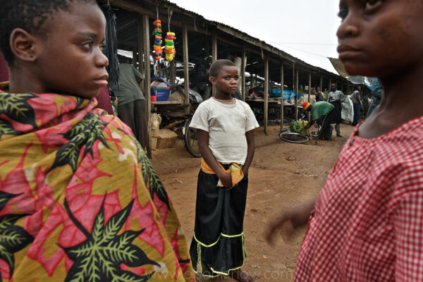 Pygmy Girls in Salate Market Feel Uncomfortable Outside the Forest
From the National Geographic story by Paul Salopek:
More than three million people have died in Congo’s six years of civil strife. This dead consists mainly of civilians. They perished mostly from starvation and disease: the worst human calamity since World War II. Yet, inevitably, it is Congo’s lurid tales of cannibalism, its sensational stories of human sacrifice, its ornamental killings, which end up bubbling into the news.
Magical violence makes it easy for journalists to reach for Joseph Conrad’s bleak fable Heart of Darkness every time a Congo headline is required. This fixation on “unspeakable” rites in “Darkest Africa” obscures the actual origins of the war: bitter ethnic grudges, meddling by powerful neighbors such as Uganda and Rwanda, and endless squabbles over Congo’s immense storehouse of gold, diamonds, coltan, and timber.
Still, this much is true: the miasma of juju is inescapable in Congo. It is like swamp vapor. Invisible. Pervasive. Soccer teams hire sorcerers to hex their rivals. Prostitutes pay good money for charms that make them irresistible. And in the nation’s Wild East, the magic becomes explosive, toxic, like the volcanic gases that are trapped in the bottom of its deepest lakes.
From my journal:
The Pygmy group we find in Beni is interesting because they have given up on the nomadic leaf structures of traditional pygmies and they have built mud and wattle houses like the Bantus…
I had sent Paluku out to scout for Pygmy groups without a forest and Pygmies to find Pygmies in Beni.  Paluku found three Pygmies working in Beni under slave like conditions.
Each time we go outside Beni it seems the soldier buildup is increasing.  We always leave early, so we aren’t hassled… But always on the return trip some thug soldier that has to get to Beni commandeers our car… They don’t give us an option… I guess it’s better than what they are doing with the Toleca drivers… they are just stealing their bicycles for the war effort.  I have to be careful though, because I have absolutely no authority to work outside the reserves.  And the commandant said straight up he didn’t like white people… His concern, I believe, is he thought they were probably all spies… The cameras stay in the backpack for the entire trip back.
Paluku decided not to tell me about the fighting in Goma, Bukavu and South Kivu so I would not be unnecessarily concerned… But he does decide it’s not a good idea for me to take moto taxis or walk around Beni anymore.  The first Kabila financed his war by kidnapping a wealthy white guy… the ransom may be the reason he ended up running the country.  I do have a satellite phone though and Melissa read me the news the other day.  Also the airport next to the logging tycoon house I am staying in is abuzz with Antonovs coming and going… I am told they are flying in commanders to rally the troops… to get them ready for war.
