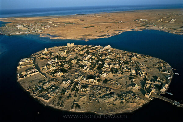 Suakin Port | Ancient, Roman and Unused | Port Sudan | Northern Sudan
The greatest asset the North has is access to the sea.  This is the old port—Suakin.  Oil goes out of a newer port. Pilgrims from all of West Africa gathered at Suakin to cross Red Sea to Mecca. The trip to this port determined settlement patterns all across the Sudan.
Suakin was ottoman built but has Roman remnants and was possibly chipped into this perfectly round circle by the Romans.  Suakin was the main port from the 14th century until World War I and has never been excavated. I was jerked out of the air right after shooting this photograph and never got up to shoot aerials again…
From my journal:
The next day our charter plane lands around noon and we fly over dust for 3 hours.  I make sure there is a case of water and the sat phone has charged batteries in case we have a forced landing.  This plane is an amalgam of parts—French body, Cessna controls and American engines.
My original plan was to just continue this flight and do aerials because we have the permissions, but a three-man crew shows up with the plane and we have three people with a lot of luggage—the plane just gets off the ground at the end of this dusty airstrip with all this weight.  I decide to land, unload and then do aerials.
We land in Port Sudan and unload and pull out some of the seats just so I can work and then wait on the tarmac.  I’m afraid if we do anything else something will get screwed up.  We take off at 5:30pm and fly south—not much to this landscape, but the light is good.  We end up circling the ancient coral port city of Suakin—it looks pretty good and the light will be perfect in half an hour.  The pilot gets a call from the control tower and is told to return immediately.  There are a lot of guns here and we don’t want to be shot down so I watch the perfect light I just paid for from the tarmac.
There is a great deal of confusion at the control tower… first they say there were no permissions circulated by Khartoum, then they say they found something but it doesn’t matter.  Cherri convenes every politician imaginable on the terrace of the Hilton—a beautiful new hotel next to the decaying government provincial building.  Most of the local ministers probably hang out here getting free drinks and food from people like us.  Cherri also calls Mahdi and miraculously his mobile phone rings in Abu Dhabi where he is attending a wedding. He calls the top general in this area from his mobile and has his office fax letters.  But it isn’t until 1am that we get the ok to fly in early morning.  Kamal forgot all his ID papers and ends up in my room because I feel sorry for him.  His snoring destroys the four hours sleep I had hoped for and when I go into the bathroom there is an incredibly gross oil slick in the tub and somehow he has managed to hose down the only toilet paper.  I guess he didn’t know how to use the fancy Arabic water wand next to the toilet.
There is major confusion here about what time it is.  The pilot says there is an hour difference between the here and Khartoum and the hotel people 10 feet away from each other give me two different times an hour apart and say there is no time difference between Port Sudan and Khartoum.
So through more confusion I get up for about an hour to photograph the red sea hills.  Later that day the dust storms start and I dismiss the plane and on no sleep, we head toward Suakin by ground route.
When we get to Suakin, there are more problems.  I have my green photographer card, passport and travel permits—but after waiting over an hour, it seems this isn’t enough.  We go to a higher-level security office and wait another hour.  It is now around 3pm and I have no photographs.  It turns out they left some of the papers in the car at Meroe when we got on the plane.  It’s now 4pm and they’ve finally worked it out but they need to go back to the other security office for “just 5 minutes.”  I say ok, but I need to work.  So they leave me and I’m photographing these old guy in various states of undress going into the ocean, their Jellabia’s blowing in the wind.   They think the waters here have healing properties.  I work for ten minutes and a pickup truck pulls up and a hulking Idi Amin type jerks my cameras out of my hands and just stands there with this stupid expression on his face.  There is nothing I can do—I ask him “are you stealing my cameras?”  “Do you just want to hold it?  What?”  After 20 minutes of grilling in Arabic by some other goon asshole, Kamal shows up and somehow smoothes things over.  I’ve spent a lot of money in the last few days and don’t have a lot to show for it. 
Now Kamal and the group want to eat…. Is it possible to work in this country or not… I let them eat and wander off in disgust to try to photograph a little at dusk in the town—later they scramble everywhere to try to find me.
As we enter the old coral city, the security goons are sitting in the dust gorging themselves on fried fish sandwiches.  They wave as we enter the narrow isthmus to the city.  A guy who lives in the rubble latches onto us and becomes an impromptu guide.  At the end of the tour, he scrambles over a big pile of coral rubble that was once a building and comes back a sack of coral to sell.  It looks like he just knocked it off the reef, but maybe it is just pieces of these fallen down buildings.
As we leave we pass the security guys again—now they are lying in the dust recovering from their huge meal—they all wave.
There’s a businessman from Jordan I keep bumping into at the hotel—he is investing in the port.  He says the Arab world is coming here in droves to invest, now that things are stabilizing a bit—the Arab world is incredibly conservative with their investments and wouldn’t be doing this if peace weren’t in the offing. Sudan has great resources.  Saudi banned the import of meat from Europe and just had 600,000 sheep shipped in for Eid. Sudan has 140 million head of free-range stock.  Only ten percent of the animals here are used domestically and only another ten percent is used for export.  They have water and large schemes for agriculture and now with oil coming on line, he thinks this is a dirt-cheap place to invest.  The people here are so poor though—the government taxes a guy with a few pots, a charcoal burner, a hut and a couple of sheep. 75 percent of Sudan is technically nomadic.
All morning is spent obtaining the permissions to go to Sinkat and Erkaweit.  Both of these areas have been designated as tourist spots by the government.
We drive two hours, drop the cook at the house we are renting and pick up Ali, who is the big man of Sinkat.  Ali takes us to yet another security office where we wait for yet another hour and then are finally ushered into the main security guy’s office.  He asks me in booming perfect English how I like America.  I tell him it is a very EASY place to live.  He says “Just like here?” with a booming laugh. 
So I have a one-hour window left tonight and then tomorrow to photograph the largest nomad group in Sudan—Beja people.
When we arrive at the market the people don’t smile or pay any attention to the camera.  This is completely different from the rest of my experience in Sudan where I am usually completely surrounded if I stand still too long.  This Beja tribe is Ethiopian and they spend most of their day making and consuming coffee in elaborate ritual just like Ethiopians.
Somehow someone in the group has talked to them about doing a very elaborate mock swordfight—probably Ali—part of their fun is to make mock assaults on me and the camera.  On the drive back to Sinkat, we stop and photograph some Hadenbowa nomads.  We are staying at Halim’s home and the cook has a meal when we arrive.  They say there are no mosquitoes here, but I see them buzzing everywhere so we put up bug nets and eat our meal.
In the middle of the night (1am or so), Kamal walks into the room and says “Oh… Randy, I am sorry, but the men from security are here and they want to take all of your cameras and films.”
