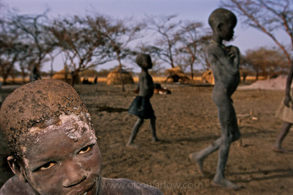 War Torn Sudan | Boys Covered in Mud Only Have Leaves to Eat
Living in the dust – In war-torn southern Sudan, boys who have only leaves to eat pack mud onto their hair to kill lice.
From the journal:
In the middle of the night (1am or so), Kamal walks into the room and says “Oh… Randy, I am sorry, but the men from security are here and they want to take all of your cameras and films.”
I’ve been asleep for 3 to 4 hours and this just seems like a bad dream—I don’t even believe him at first so I don’t make any motions to crawl out of my bug net cocoon.
Out of the cocoon, it’s cold, reality is setting in and both facts have me physically shaking.  I am carrying one third of the total film I’ve shot so far and that includes $5,000 in aerials—not to mention expensive camera equipment in the hands of security goons.  These goons are the kind of raw material they hire at the Hilton and they have to teach them what a fork is before they can even let them bus tables.
We make multiple phone calls to our security guy in Khartoum and he knows nothing about this.
The goons won’t even tell us their names or show any form of ID.  They just say they are giving us this one chance to turn over all the materials and that this order comes from Khartoum.  They say they are very sorry, but it is out of their control—they won’t even talk to our assigned security agent—Anwar–in Khartoum on the phone.  They say their orders come from Imad-Head of Internal Security for Port Sudan.  Anwar calls Imad and finds that Imad never gave any such order—but it’s too late, they’ve taken the stuff after a veiled threat about taking us to jail.  The booming guy says “take it easy, this is life,” as he leaves with my pelican case in his right hand.
Until 1998 security goons showed up like this in the middle of the night and people just disappeared.  This even happened as late as 2001 with Turabi’s people.
I try to go back to sleep, but it isn’t working, I am too angry, my heart is pounding too hard. I get out the sat phone to try to call somebody, but I don’t even know who to call.  I’m thinking what press organization could use this abuse as a nut graph in a story about this police state.
Cherri gives me half an Ambien and I get about an hour of sleep.
We pack up in the morning—grab bread, boiled eggs and jam (they insist on buying jam because they think “Americans want jam.”).  Ali, our local guide, who was also here in the middle of the night with the Gestapo, looks like a bus hit him.  He is so sad for us.  A cell phone call to Imad tells us to not even come by the local security office, that my equipment was put on a plane to Khartoum and if I want it that I should just follow it back.  In other words telling us to get the hell out of town.  Why are they taking my stuff to Khartoum if no one in Khartoum ordered it?  We wonder if there is an imminent rebel attack and this is all a ruse to get us out of the area.  Mahdi used a similar tactic to get Cherri’s group out of an area that could get hot.
So we rearrange stuff in the car, not knowing if we’ll be searched at the airport or not.
I had a 30 second window away from the goons and managed to separate all my shot film, notes, expense book, and malaria tabs out of the main case I gave them while Cherri kept the goons busy last night.  I hid this in Cherri’s underwear in her suitcase; figuring Islamic goons would be embarrassed to look there.  Now we are too afraid to carry the film through the airport.  Sammy—the driver–pulls over and I sort out the film and cover it with sleeping bags.  We line our bags of clothes up on top so we can just grab them and Sammy can speed off driving my film back to Khartoum.
Kamal has been trying to call his cousin who is head of security for this airport.  It’s probably appropriate to mention here that Osama Bin Laden built this airport.
We meet the cousin at the front door of the airport and he tells Kamal that there are three security guys waiting to secretly accompany us on this plane.
I have no idea what will be waiting for us in Khartoum.  Maybe a more fundamentalist group in government is behind this and we will be stopped at the Khartoum airport.  The weirdest part of this is just not knowing what is going on or why.  We know security is tight in Port Sudan ever since the U.S. launched cruise missiles from the Red Sea to blow up the Shifa pharmaceutical company and security was reamed for not seeing it coming.  So these folks are not particularly pro-American… And when they see me in a plane circling and circling and taking pictures… even though they are told I have all the clearances to do this, they just don’t believe it… no one has been allowed to do this in twenty years.
Yahia Babiker is in charge of all external security in the country and we finally get through to him.   He says my equipment was hand carried by an agent this morning but then when we land in Khartoum we are told my equipment and film will come with someone on an evening flight.
We have a meeting with Yahia at 3pm, but he doesn’t want to meet until he can find my stuff.  Who knows where it is or what is going on.  We are told Mahdi and Yahia are mad about this and will fire people involved—I doubt it—They can’t control them from Khartoum, how will they fire them out of their little fiefdoms if they weren’t even aware of them in the first place?
Yahia’s office is actually a compound with many cars and drivers and security guys everywhere within this little walled city.  This is Sudan’s version of the KGB—there isn’t the confusion here that reigns in the other government camps.  The guy at the gate looks at us and knows where we are going.
Yahia’s office is new, clean and sparse—very bright cool white fluorescents overhead with modern diffraction grids in a drop ceiling—but it’s a new drop ceiling and the fixtures aren’t whining as they are in the rest of this country.  There is a grey-blue leather seating area in an L-shaped office.  The other side of the L is Yahia’s desk with the latest computer equipment (i.e. COMPUSA new, nothing fancy).  The only other item in this large room is water cooler on the slate blue floor.  We are sitting in the chairs of the leather rap pit and he is on the big sofa with only his cell phone.  The ministers talking on their tiny cell phones run this entire government.  We asked Yahia for his cell phone number, but he refused.  The cell phone rings periodically—he squints at the number and then pushes a button to get rid of the call.  These guys all sit around and wait for the number to show up as Bashir.  I can’t tell if this is a scary man or not.  I know Yahia’s predecessor had “ghost houses”  all over Khartoum where people were tortured.  So, in a stupid move, I invade his space, sit close to him on the couch and show him a copy of NG that has a story by Paul and a story by me.  He responds fairly well so I stay there.  He says that for our “inconvenience” he has tried to make up for it by spending the morning clearing our trip to the oil fields.
As the meeting ends, Paul asks him about the University of Montana and Yahia says he went to the Univ. of Missouri.  I tell him I taught there in the Journalism School and he brings up all sorts of names of professors—this conversation is surreal.  All the ministers I’ve met have gone to Midwest party schools.  Yahia majored in political science at MU which I know is not much of a program—and who knows if he was even in the top half of his class.  Yahia and I missed being at MU at the same time by one year.  We have former students sitting in fotohut booths in grocery store parking lots with similar academic records as the folks that are running this country.  And what is really scary is that the next level of government below the ministers is like falling off a cliff into imbecile land.
Yahia takes us into the next room where they have my case with most of the cameras, but no film or videotapes.  One of the cameras is flashing numbers, indicating that film was ripped out of it.  I explain a Leica and 35mm lens are missing (about $5000) and point to the space in the case where it used to be.  Yahia just asks if it was insured.
