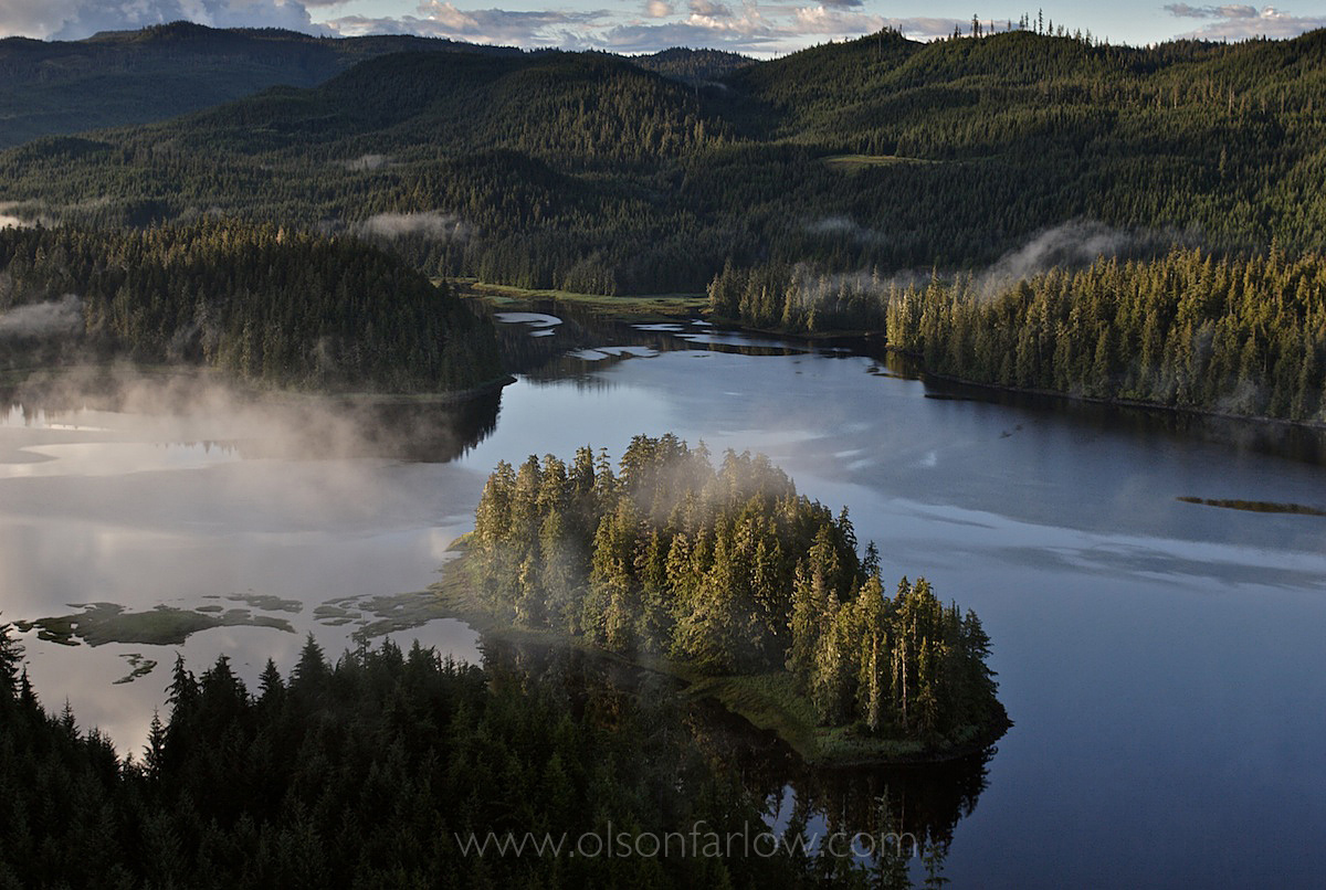 Bird's eye view of tiny island surrounded by water and fog rising from surrounding landmass.